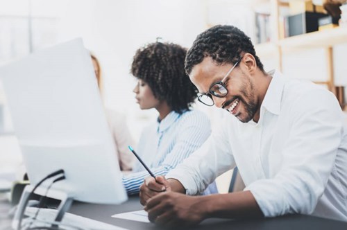 man writing notes sat in front of computer screen 