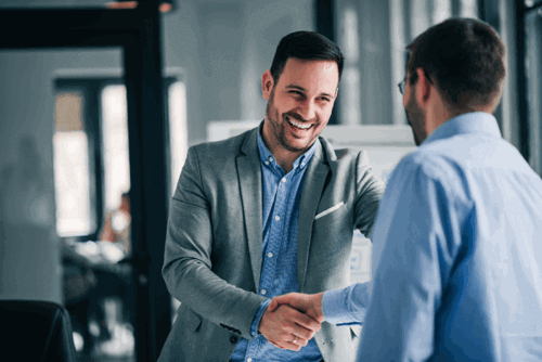 Two professionally dressed men smiling and shaking hands in a modern office setting, suggesting a successful business meeting or agreement.